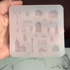 May include: A clear, square silicone mould with a miniature town design. The mould includes detailed shapes of houses, trees, street lamps, a church, and a park bench. The text "MINI DESIGN" is visible at the bottom.