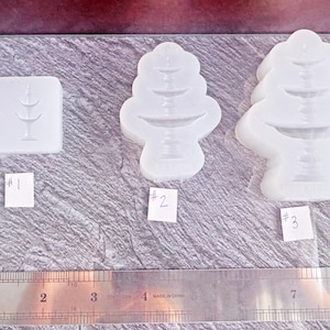 Puede incluir: Tres moldes de silicona transparentes para bandejas de servicio en capas. Los moldes tienen diferentes formas, incluido un molde rectangular con dos bandejas y dos moldes en forma de árbol. Una regla se coloca debajo para referencia de tamaño.