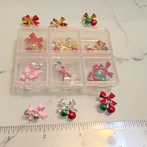 Può includere: Un organizer di plastica trasparente pieno di vari ciondoli a tema natalizio. I ciondoli includono fiocchi rossi e dorati, ornamenti verdi e rossi e fiocchi rosa. Un righello è posto sotto, misurando in centimetri.