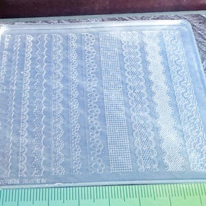 May include: A clear, rectangular nail art stamping plate with diverse lace patterns. The plate is labelled with "Kusa collection" and "Gingin". A green ruler is at the bottom, and the plate is approximately 10 cm wide.