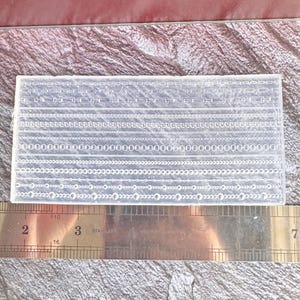 Puede incluir: Un molde de silicona transparente y rectangular con varios diseños de cadenas y eslabones. Una regla metálica con marcas de centímetros se coloca junto al molde. El fondo es una superficie texturizada de color granate.