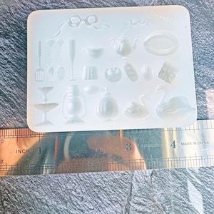 May include: A clear silicone mold with various miniature shapes, including a teapot, swan, and gift box. Other shapes include a champagne flute, spoon, and a small plate. The mold is approximately 4 inches wide.