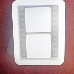 May include: A clear, rectangular silicone mold with rounded corners. The mold features two square spaces and a film strip design along the sides. The mold is designed for crafting and creating resin art or similar projects.