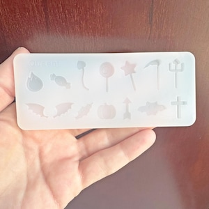 May include: A clear silicone mold with various Halloween-themed shapes, including a candy corn, a lollipop, a star wand, a scythe, and a trident. The mold also features a bat, a leaf, a pumpkin, an arrow, a cloud, and a cross. The word "Queeni" is printed on the mold.