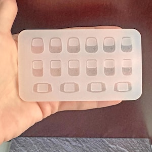 May include: A clear, rectangular silicone mold with 15 small, rounded rectangular cavities. The mold is held in a hand, showcasing its translucent quality and the detailed shape of each cavity, ideal for crafting small items.