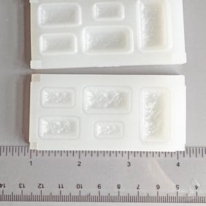 Peut inclure: Deux moules en silicone blancs avec des cavités rectangulaires et carrées. Une règle est placée en dessous, indiquant des mesures en centimètres. Les moules sont probablement pour la création d'objets rectangulaires.