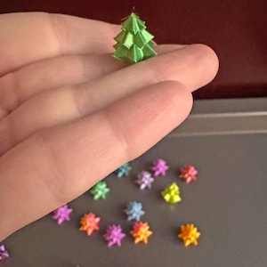 May include: A miniature green Christmas tree made of folded ribbon sits on a hand. Scattered around are colorful star-shaped ornaments in shades of pink, orange, blue, and yellow. The background is a dark red.