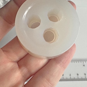 May include: A circular, off-white object featuring three circular holes. The object appears to be made of a translucent material. The item is held in a hand, with a ruler in the background.