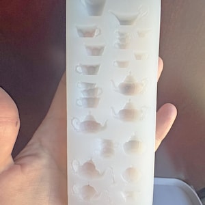 Puede incluir: Un molde de silicona transparente y rectangular con tazas de té, teteras y una jarra de crema en miniatura. El molde se sostiene en una mano y está diseñado para crear pequeños objetos decorativos. El diseño incluye varias formas y tamaños.