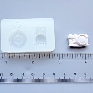 May include: A clear silicone mold with two designs: a pocket watch and a camera. A small, unpainted camera is next to the mold. The mold is placed on a ruler, with measurements in inches. The camera is approximately 1 inch long.