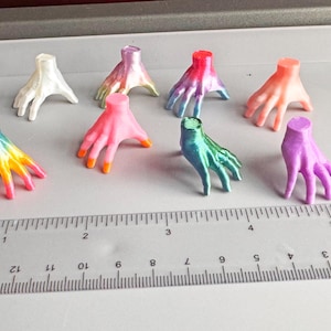 Puede incluir: Una colección de ocho esculturas de manos en miniatura en varios colores, incluyendo blanco, rosa, naranja, verde y morado. Las manos están dispuestas sobre una superficie gris junto a una regla, mostrando su pequeño tamaño y detalle.