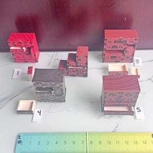 Könnte beinhalten: Eine Sammlung von fünf Miniatur-Holzkommoden mit verzierten Metallbeschlägen, beschriftet von 1 bis 5. Die Kommoden haben rote oder dunkle Holzoberflächen und herausnehmbare Schubladen. Ein 15-cm-Lineal liegt unten als Maßstab dabei.