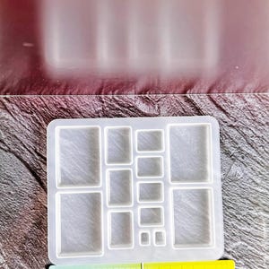 May include: A clear silicone mould with various rectangular and square cavities. A ruler with centimetre measurements is placed below the mould. The mould is designed for crafting projects, such as resin art or jewellery making.