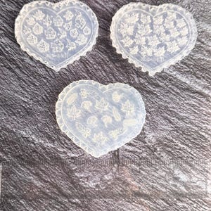 May include: Three clear silicone molds in a heart shape, featuring leaf designs. The molds have a decorative scalloped edge and the text "mo-nail" is visible on each. The molds are arranged on a dark, textured surface.