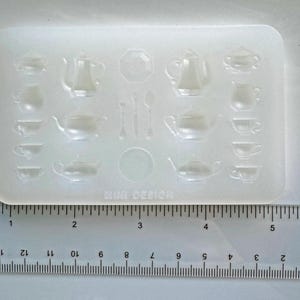 May include: A clear, rectangular silicone mold featuring miniature tea sets. The mold includes teapots, cups, saucers, spoons, and a sugar bowl design. The text "MINI DESIGN" is printed at the bottom. A ruler is placed below the mold.