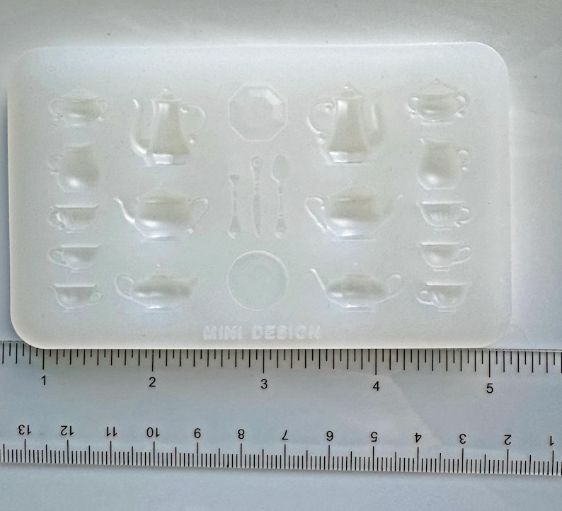 May include: A clear, rectangular silicone mold featuring miniature tea sets. The mold includes teapots, cups, saucers, spoons, and a sugar bowl design. The text "MINI DESIGN" is printed at the bottom. A ruler is placed below the mold.