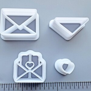 May include: Four white plastic cookie cutters in the shapes of envelopes and hearts. One envelope is square, another is triangular, and the third has a heart cutout. The fourth cutter is a heart shape.
