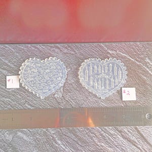 May include: Two clear, heart-shaped molds with scalloped edges. One mold features a design of swirling patterns, and the other depicts candles. A ruler with inch measurements is placed below the molds. Small labels with the numbers #1 and #2 are visible.