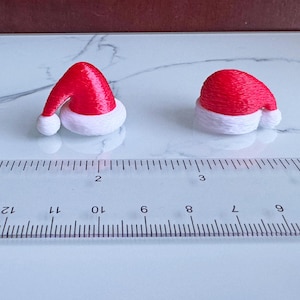 Könnte beinhalten: Zwei Miniatur-Weihnachtsmützen. Die Mützen sind rot mit weißem Besatz und einem weißen Bommel. Die Mützen sind auf einer weiß-grauen Oberfläche neben einem Lineal platziert. Das Lineal zeigt Messungen in Zentimetern an.