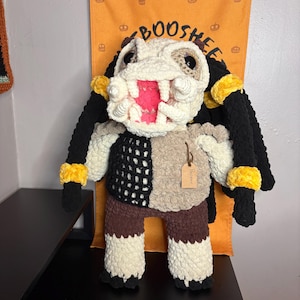 May include: A handmade crochet plush figure featuring a cream-colored head with a wide, open mouth revealing pink interior details, large black eyes, and long black dreadlocks with yellow bands. The body has a black and tan torso and brown legs.