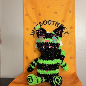 May include: A crocheted plush cat with black and neon green stripes and multicolored speckles. It features mismatched button eyes, a stitched pink nose, and white fangs. The cat is positioned in front of an orange fabric banner with pumpkin icons and the text JUST BOOSHET.