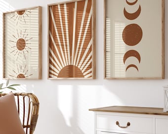 May include: Three framed prints with abstract sun and moon designs in shades of brown and beige, hanging on a white wall above a white cabinet with a wooden top. A wicker chair with a brown cushion and a white throw blanket sits in front of the cabinet.