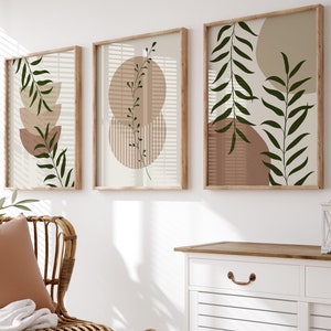 Puede incluir: Tres impresiones de arte abstracto enmarcadas con una paleta de colores neutros beige, marrón y verde. Las impresiones presentan hojas estilizadas y formas geométricas. Las impresiones están colgadas en una pared blanca sobre una cómoda blanca con dos cajones.