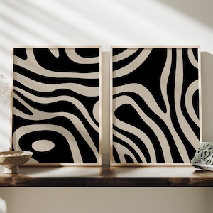 Op de afbeelding: Twee ingelijste abstracte kunstprints. Elke print heeft een zwart en beige golvend patroon. De frames hebben een lichte houtkleur. De prints worden op een donkere houten tafel getoond.