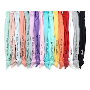 Pu&ograve; includere: Una collezione di quindici magliette disposte fianco a fianco in uno spettro di colori che include Heather Prism Mint, Peach, Ice Blue, Sunset, Dusty Blue, Lilac, Team Purple, Pink, Sea Green, Mint, Yellow, Orange, Red, White, Athletic Heather e Black.