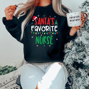 May include: A black sweatshirt with a Christmas-themed design that says "Santa's Favourite Nurse" in red, green, and yellow letters. The design also includes a Santa hat, a Christmas tree, and a string of Christmas lights.