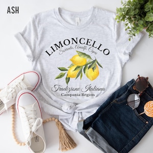 Pu&ograve; includere: Una t-shirt grigio melange chiaro con una grafica di tre limoni gialli con foglie verdi. Il testo recita LIMONCELLO, Sorrento, Amalfi, Capri, Tradizione Italiana e Campania Region in caratteri neri, stilizzati e con grazie.