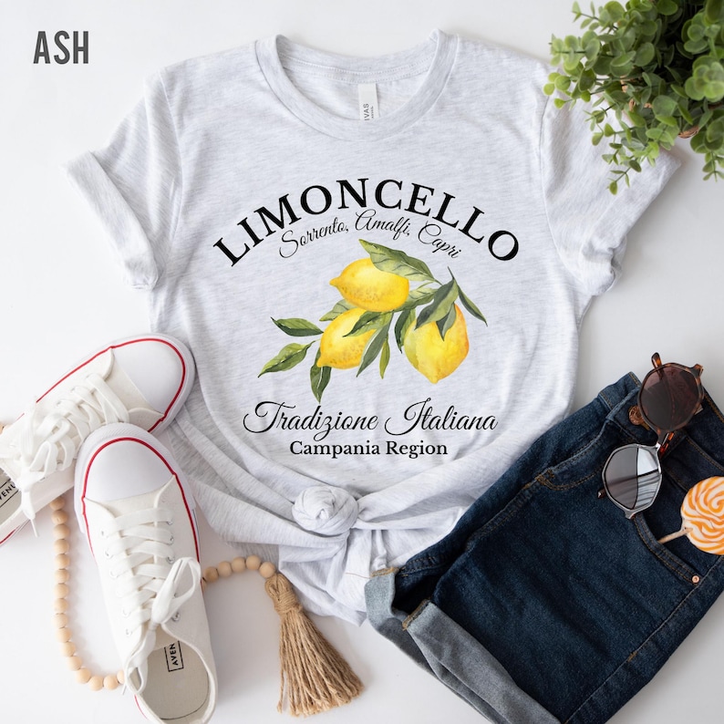 Pu&ograve; includere: Una t-shirt grigio melange chiaro con una grafica di tre limoni gialli con foglie verdi. Il testo recita LIMONCELLO, Sorrento, Amalfi, Capri, Tradizione Italiana e Campania Region in caratteri neri, stilizzati e con grazie.
