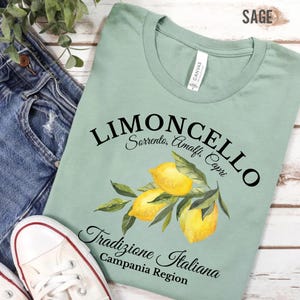 Pu&ograve; includere: Una t-shirt verde salvia con una grafica di tre limoni gialli con foglie verdi. Il testo riporta LIMONCELLO, Sorrento, Amalfi, Capri, Tradizione Italiana e Campania Region in caratteri neri. La t-shirt presenta un girocollo.