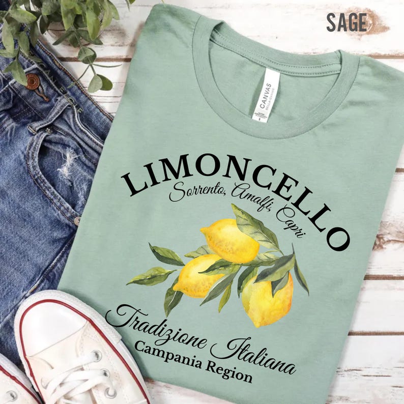 Pu&ograve; includere: Una t-shirt verde salvia con una grafica di tre limoni gialli con foglie verdi. Il testo riporta LIMONCELLO, Sorrento, Amalfi, Capri, Tradizione Italiana e Campania Region in caratteri neri. La t-shirt presenta un girocollo.