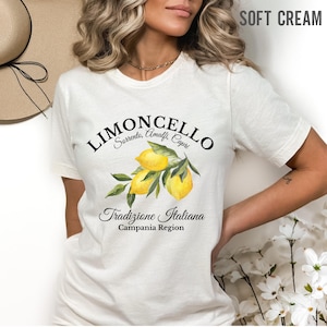 Pu&ograve; includere: Una t-shirt a maniche corte color crema tenue con una grafica di tre limoni gialli con foglie verdi. Il testo riporta Limoncello, Sorrento, Amalfi, Capri, Tradizione Italiana e Campania Region in un carattere nero.