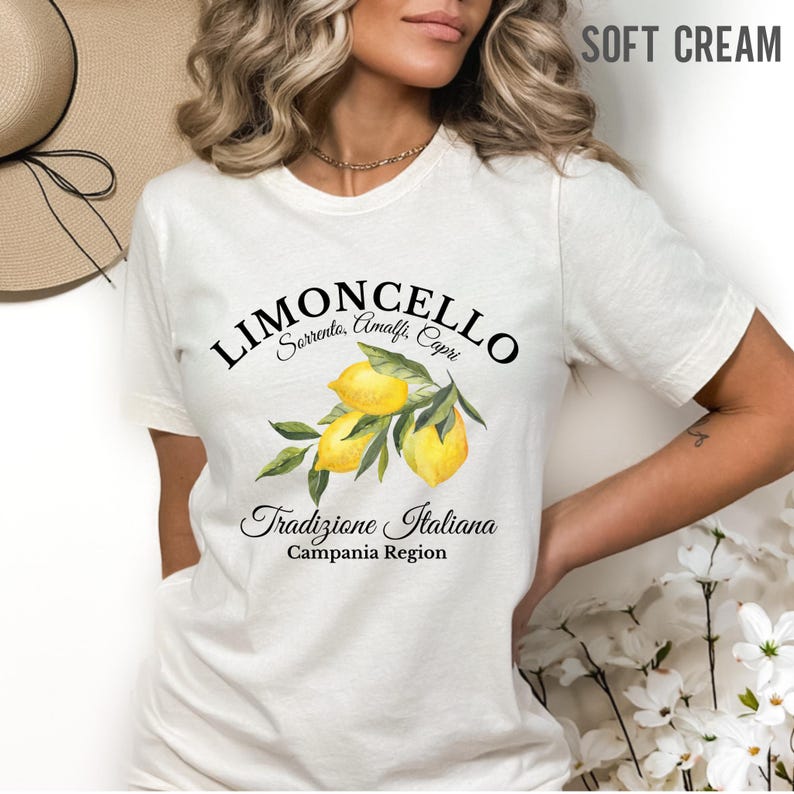 Pu&ograve; includere: Una t-shirt a maniche corte color crema tenue con una grafica di tre limoni gialli con foglie verdi. Il testo riporta Limoncello, Sorrento, Amalfi, Capri, Tradizione Italiana e Campania Region in un carattere nero.