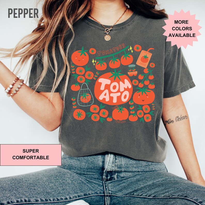 May include: A dark gray t-shirt with a tomato graphic. The graphic features a large tomato with the word "TOMATO" written on it, surrounded by smaller tomatoes. The text "TOMATOES" is written above the large tomato. The shirt has a crew neck and short sleeves. The text "SUPER COMFORTABLE" is written on a pink rectangle in the bottom left corner of the image. The text "MORE COLORS AVAILABLE" is written on a pink starburst in the top right corner of the image.