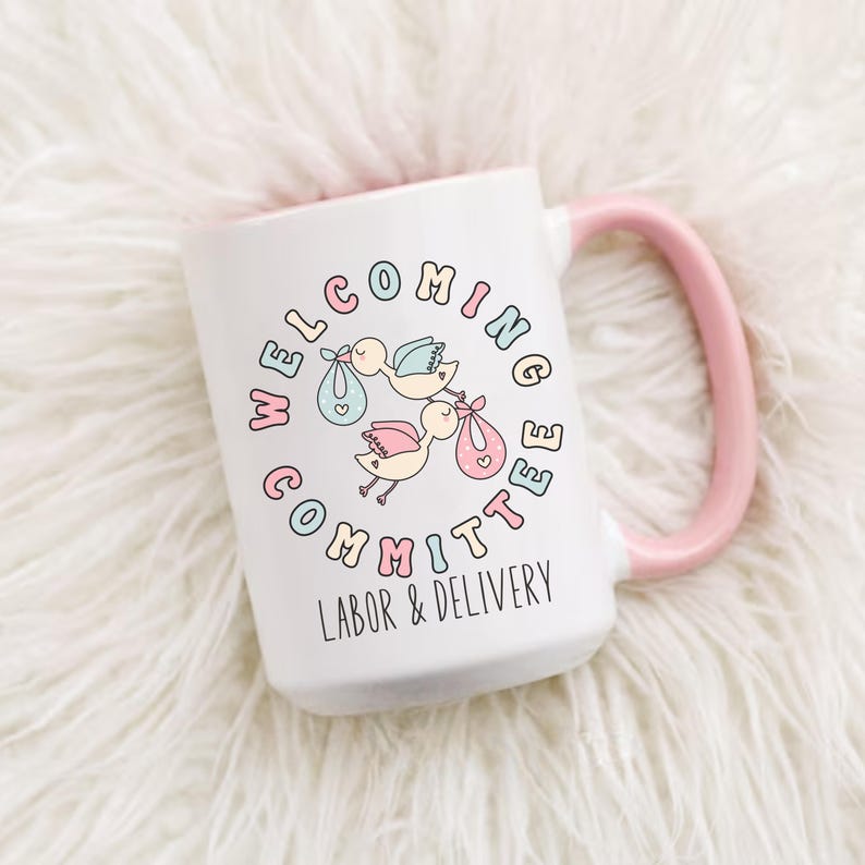 May include: White ceramic mug with a pink handle and interior. The mug features the text "WELCOMING COMMITTEE LABOR & DELIVERY" in a circle around illustrations of storks carrying babies in bundles. The storks and text are in pastel colors.