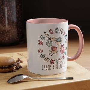 May include: White ceramic mug with a pink interior and handle. The mug features a circular design with the words "Welcoming Committee Labor & Delivery" and illustrations of storks carrying babies in bundles. A spoon and a pastry are on a wooden surface.
