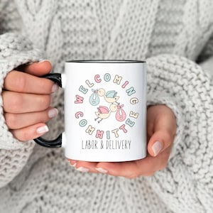 May include: White ceramic mug with a black handle and rim. The mug features the text "WELCOMING COMMITTEE LABOR & DELIVERY" in pastel colors, surrounding an illustration of storks carrying babies in bundles. The mug is held by a person wearing a gray sweater.