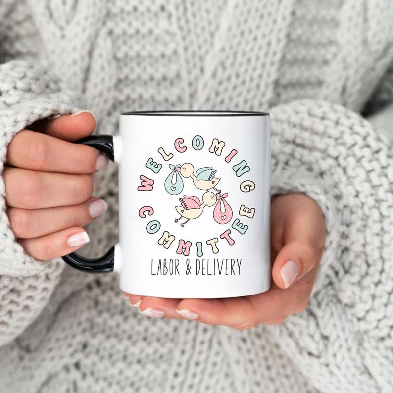 May include: White ceramic mug with a black handle and rim. The mug features the text "WELCOMING COMMITTEE LABOR & DELIVERY" in pastel colors, surrounding an illustration of storks carrying babies in bundles. The mug is held by a person wearing a gray sweater.