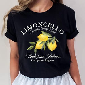 Pu&ograve; includere: Una t-shirt nera a maniche corte con una grafica di tre limoni gialli su un ramo verde con foglie. Il testo recita LIMONCELLO, Sorrento, Amalfi, Capri, Tradizione Italiana e Campania Region in caratteri bianchi e corsivi.