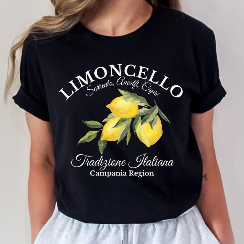 Pu&ograve; includere: Una t-shirt nera a maniche corte con una grafica di tre limoni gialli su un ramo verde con foglie. Il testo recita LIMONCELLO, Sorrento, Amalfi, Capri, Tradizione Italiana e Campania Region in caratteri bianchi e corsivi.