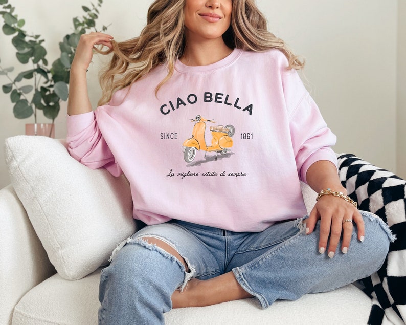 K&ouml;nnte beinhalten: Ein rosa Sweatshirt mit einem Aufdruck eines gelben Rollers und dem Text "Ciao Bella Since 1861 La migliore estate di sempre".