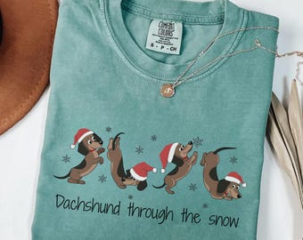 May include: Light green t-shirt featuring a graphic of dachshunds in Santa hats and snowflakes. The text "Dachshund through the snow" is printed below. The shirt has a crew neck and short sleeves.