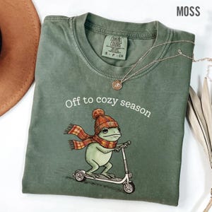 May include: A moss green t-shirt featuring a graphic of a frog wearing a knit hat and a plaid scarf while riding a scooter. The text "Off to cozy season" is printed above the frog. The shirt has a crew neckline and a Comfort Colors brand label.