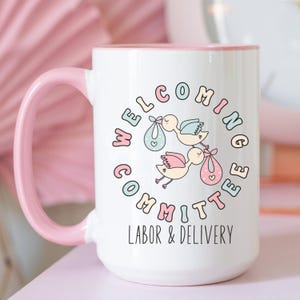 May include: White ceramic mug with a pink handle and rim. The mug features a circular design with the words "Welcoming Committee" and "Labor & Delivery". The design includes cartoon storks carrying babies.