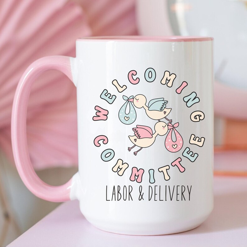 May include: White ceramic mug with a pink handle and rim. The mug features a circular design with the words "Welcoming Committee" and "Labor & Delivery". The design includes cartoon storks carrying babies.