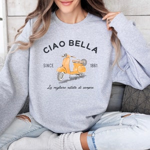 K&ouml;nnte beinhalten: Grauer Pullover mit einem gelben Roller-Motiv und dem Text "Ciao Bella" mit den Worten "Since 1861" und "La migliore estate di sempre" darunter.