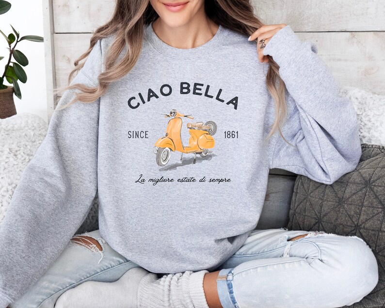K&ouml;nnte beinhalten: Grauer Pullover mit einem gelben Roller-Motiv und dem Text "Ciao Bella" mit den Worten "Since 1861" und "La migliore estate di sempre" darunter.